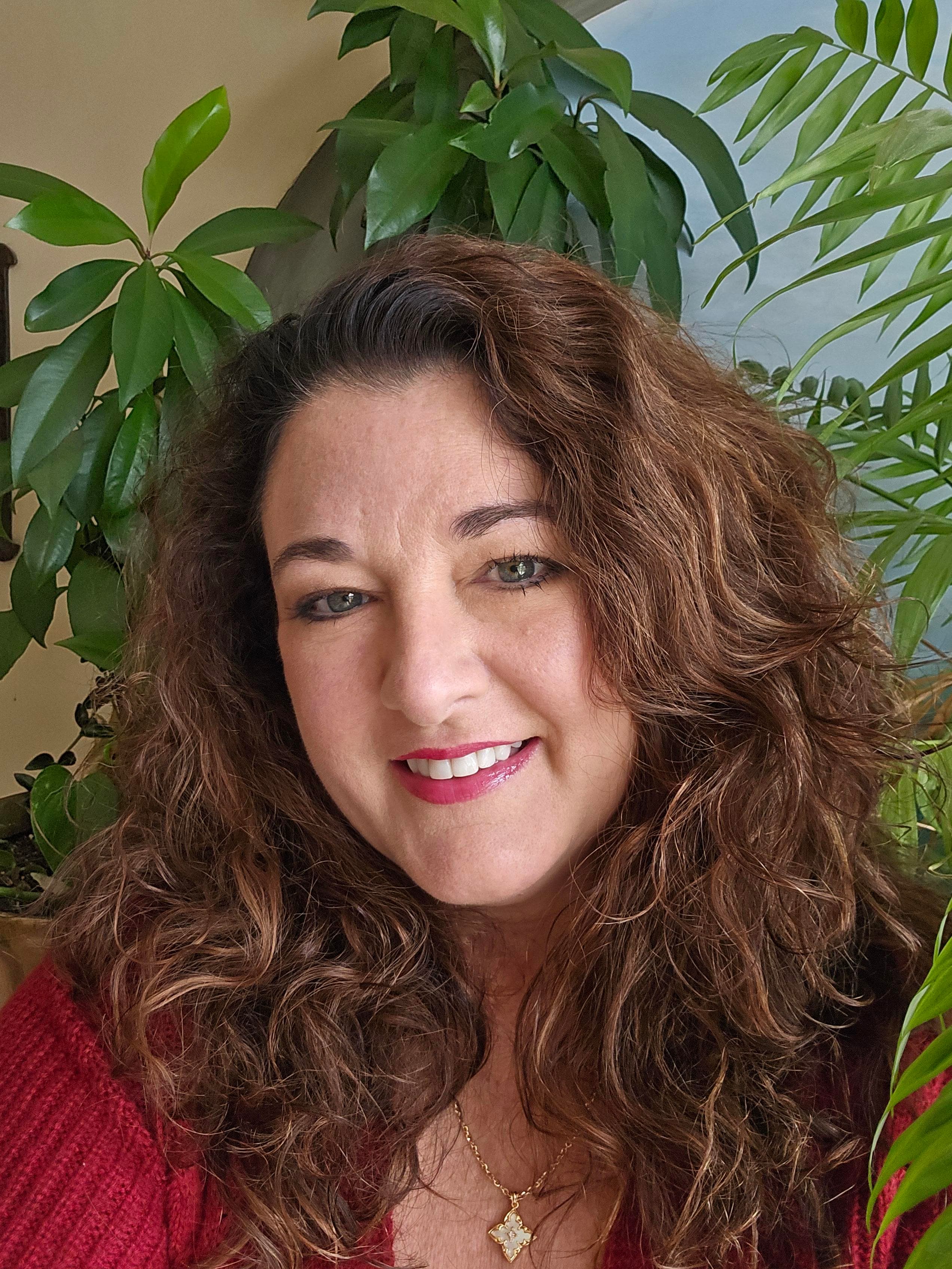 Woman with long curly brown hair and red lipstick smiles amidst lush green indoor plants.